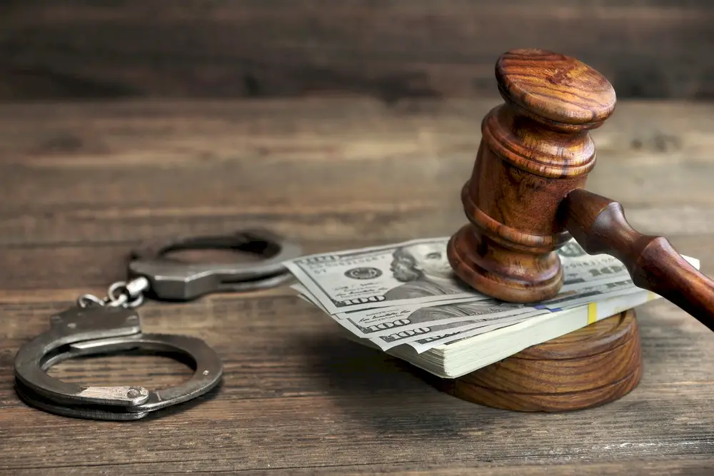 US dollar bills between gavel and light wooden table with handcuffs on left side